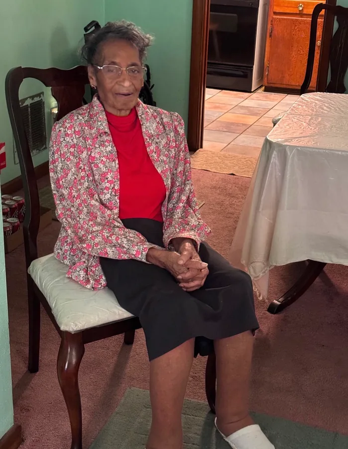 A family elder in a red blouse and patterned jacket, seated at home.