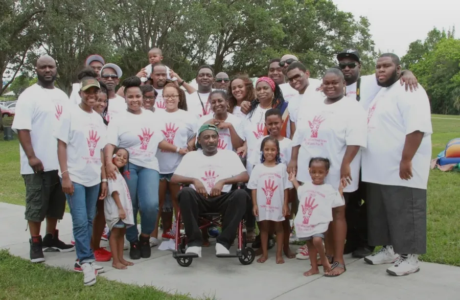 Relatives in white reunion shirts printed with a red handprint, gathered on a path under the oaks.
